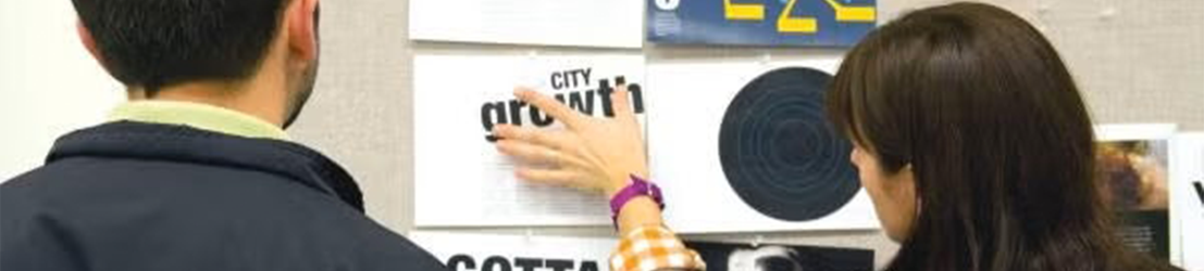 Two male students wearing white shirts writing on a transparent glass board solving a math equation in a well-lit classroom.
