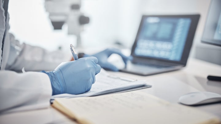 A researcher writing on a piece of paper with a pen and working on a laptop in a laboratory.