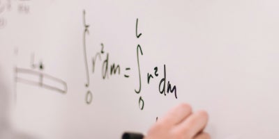 A person writing a physics formula with a marker on a whiteboard.