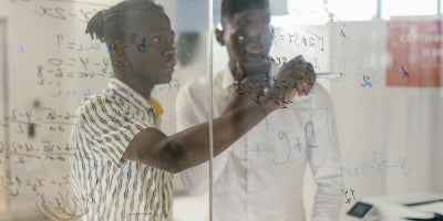 Two male students wearing white shirts writing on a transparent glass board solving a math equation in a well-lit classroom.