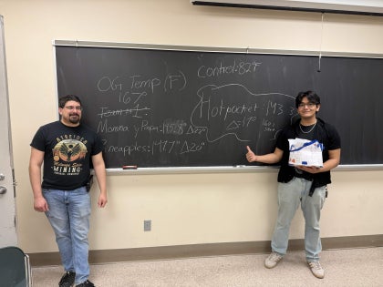 Two male students standing near a chalkboard with written formulas in a classroom.