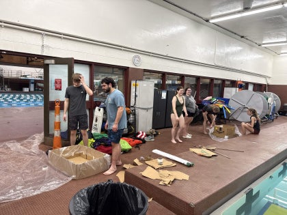 A group of students near a swimming pool with a cardboard boat and paddles.