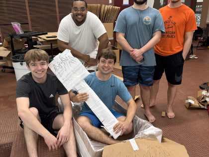 A group of students posing for a picture while holding a cardboard paddle and boat.