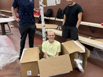 Three students posing for a picture with a cardboard boat in a swimming facility.